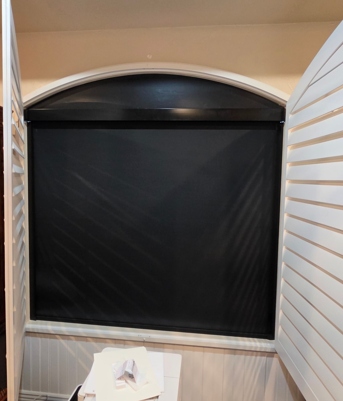 Black roller shade on an arched window flanked by plantation shutters
