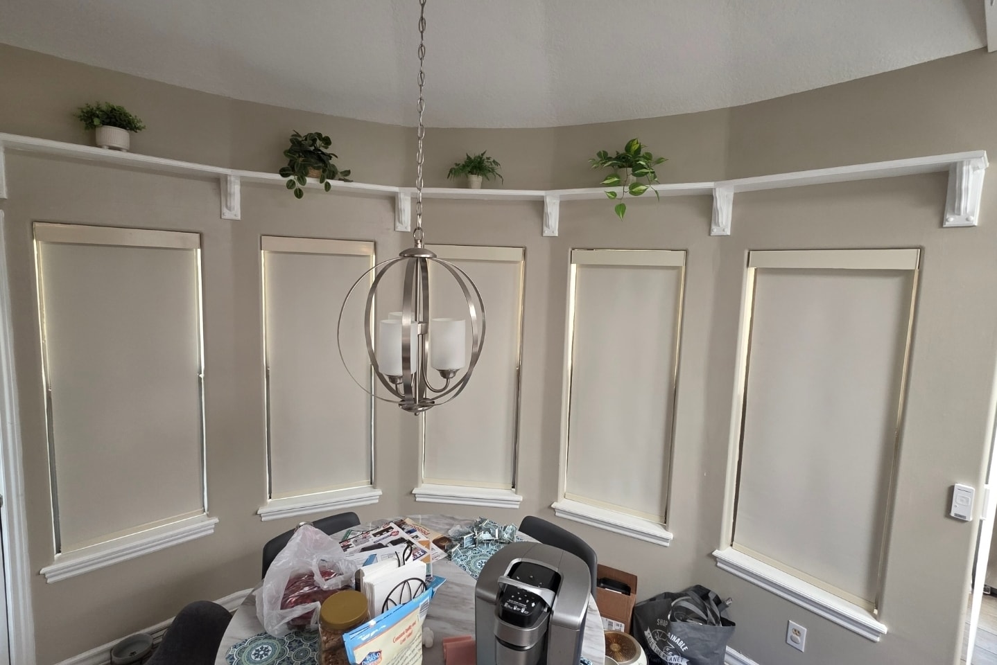 Cream roller shades wrapping a curved dining room with chandelier and plants